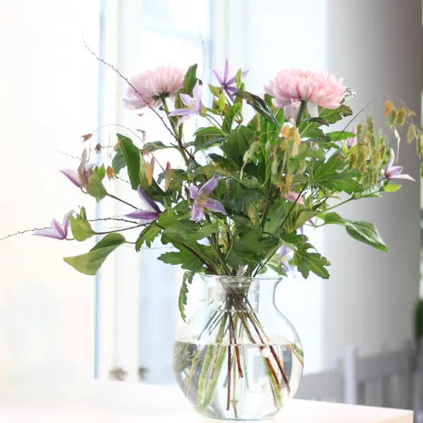 Boeket roze bloemen en blauwpaarse clematis in een bolle glazen vaas met water voor een raam. 4