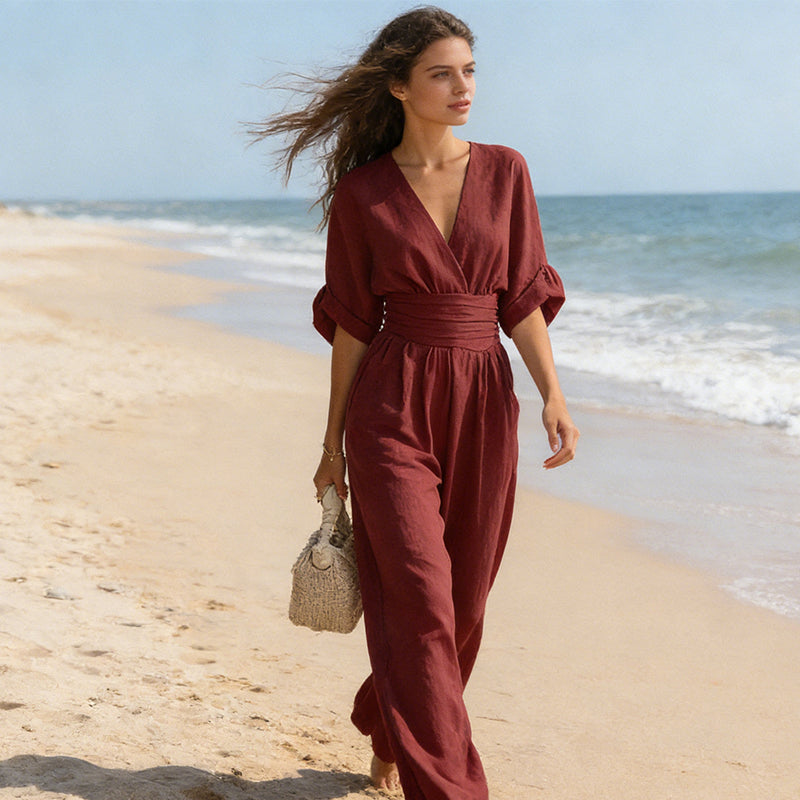 Model in wijnrode jumpsuit wandelt over het strand, met wapperend haar, de zee op de achtergrond en een rieten tasje. 7