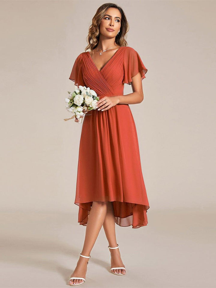 Model in terracottakleurige jurk houdt een boeket witte bloemen vast, voorkant met asymmetrische zoom en witte sandalen. 95