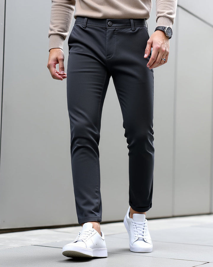 Donkergrijze chino met omgeslagen pijpen, gedragen met een beige trui en witte sneakers voor een grijze wand. 9