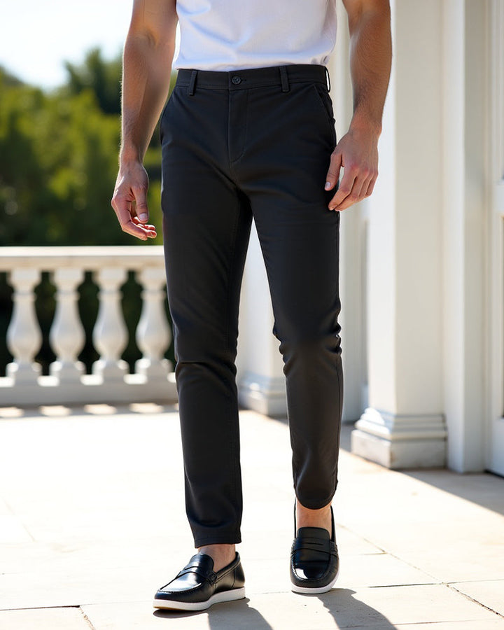 Donkergrijze chino op een zonnig terras met witte zuilen, gedragen met een wit shirt en zwarte loafers. 7