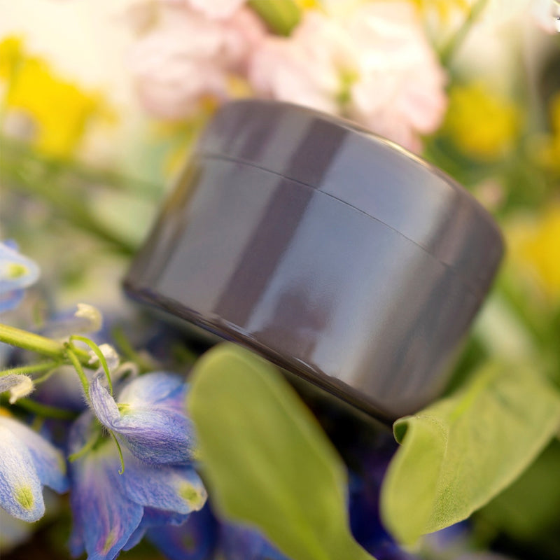 Grijze ronde inzet met deksel tussen blauwe en gele bloemen en groene bladeren, close-up met wazige achtergrond. 16