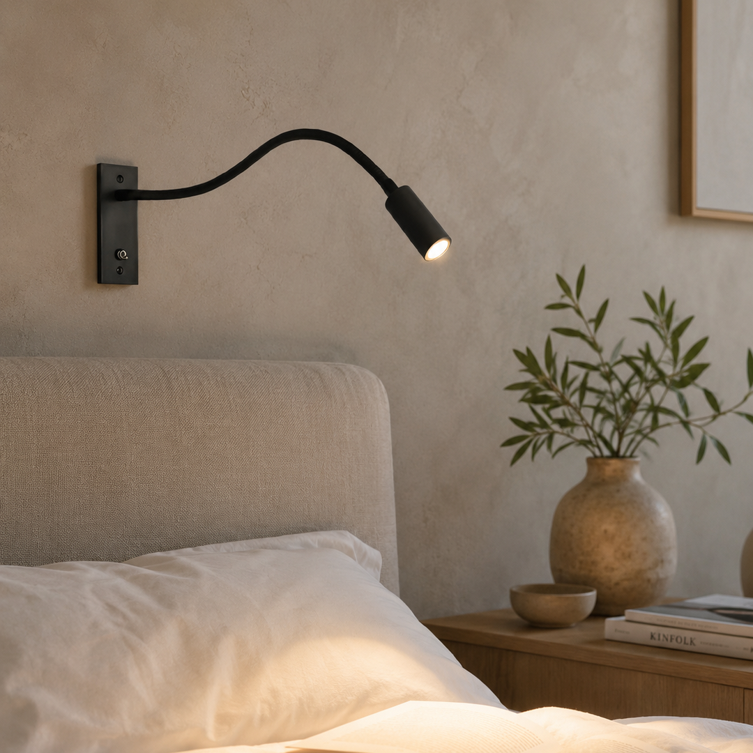 Zwarte wandlamp brandt warm boven een bed in een slaapkamer met gepleisterde muur, plant in vaas en boeken. 3