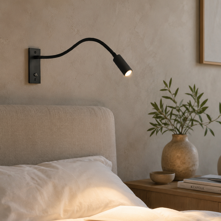 Zwarte wandlamp brandt warm boven een bed in een slaapkamer met gepleisterde muur, plant in vaas en boeken. 3