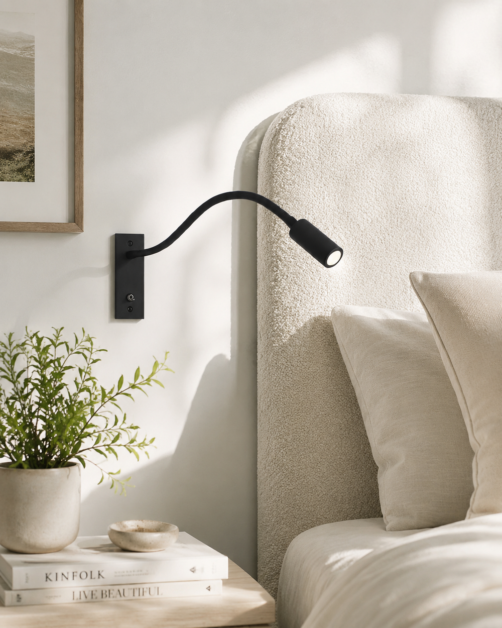 Zwarte leeslamp aan de muur naast een beklede bedhoofdeind, met nachtkastje, plant en boeken op de voorgrond. 2