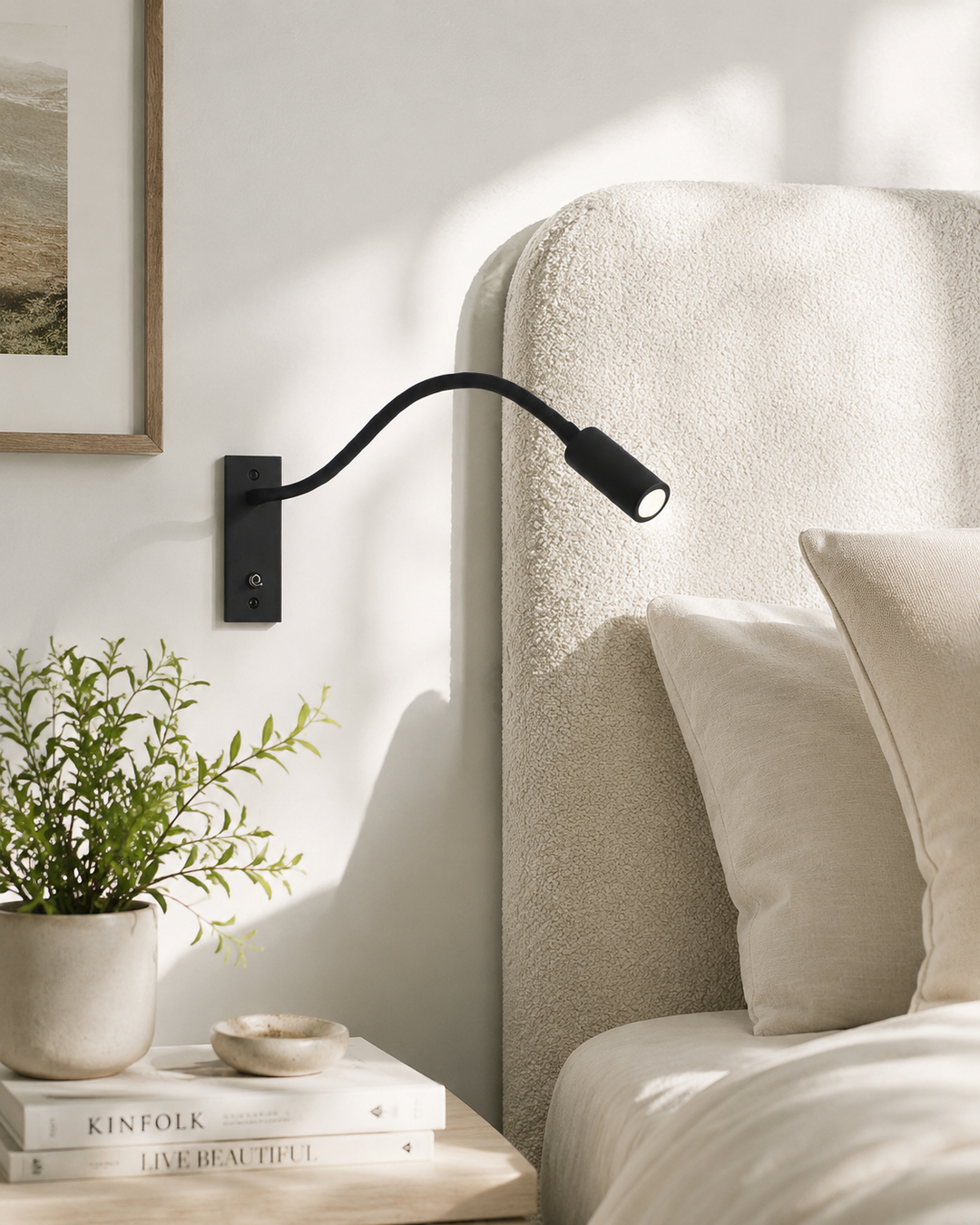 Zwarte leeslamp aan de muur naast een beklede bedhoofdeind, met nachtkastje, plant en boeken op de voorgrond. 2