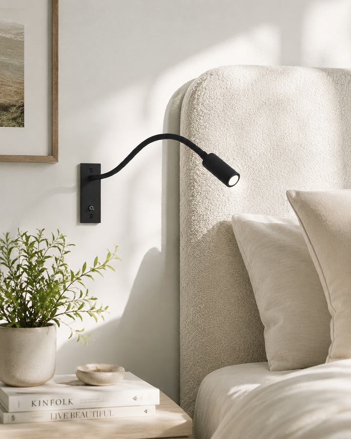 Zwarte leeslamp aan de muur naast een beklede bedhoofdeind, met nachtkastje, plant en boeken op de voorgrond. 2