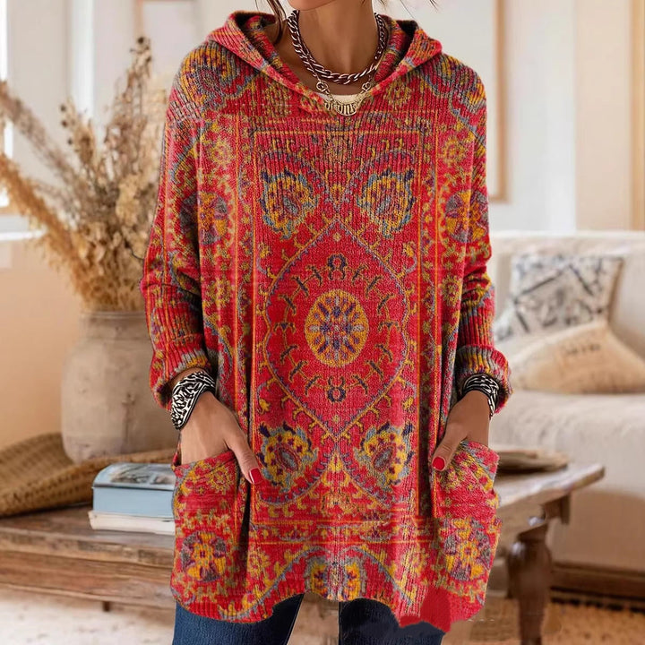 Model draagt de gebreide tuniek in rood met geel tapijtmotief, handen in de zakken, in een woonkamer met droogbloemen. 1