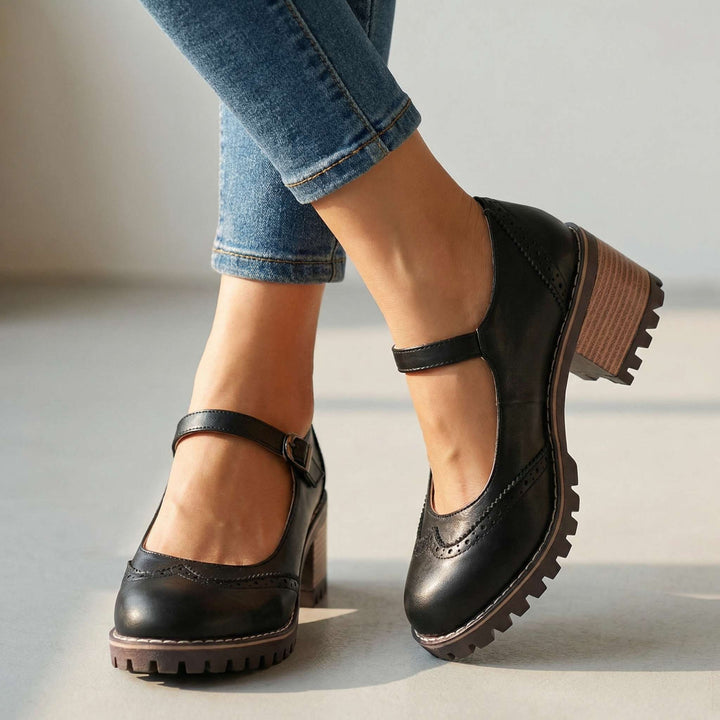 Zwarte Mary Jane-schoenen met brogue-perforatie en bandje aan de voeten van een model in spijkerbroek, close-up op effen ondergrond 1