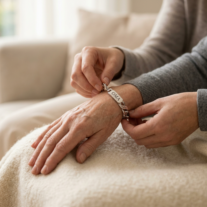 Twee handen doen een zilverkleurige armband met gegraveerd middenplaatje om de pols van een oudere vrouw, zittend op een crèmekleurige deken. 3