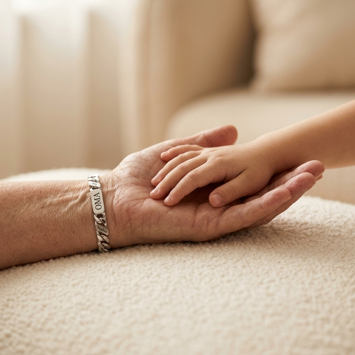 Zilverkleurige armband om de pols van een oudere hand, waarin een kinderhandje rust, op een zachte crèmekleurige ondergrond. 5