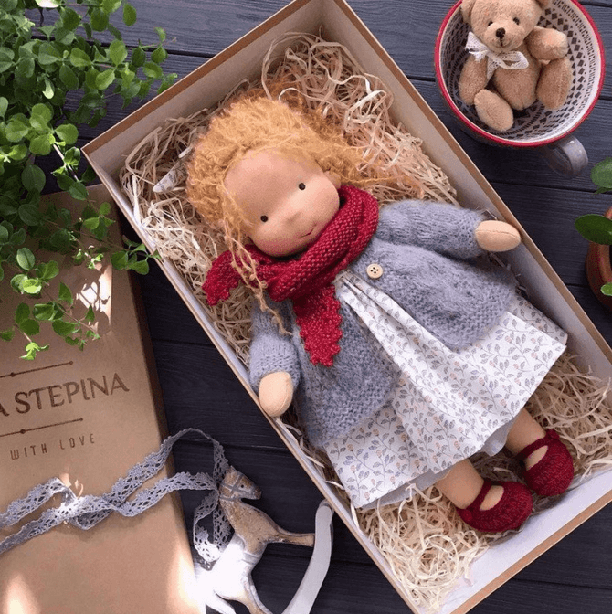 Pop met rode krullen, rode sjaal, grijs vest en jurk met bloemetjes in een doos naast een teddybeer in kopje 9