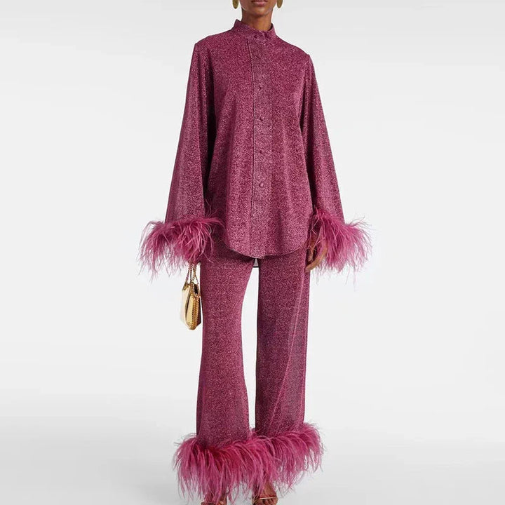 Model in bordeauxroze glitterset met knoopblouse, opstaande kraag en brede broek, beide met roze veerrand, een gouden tasje in de hand. 1
