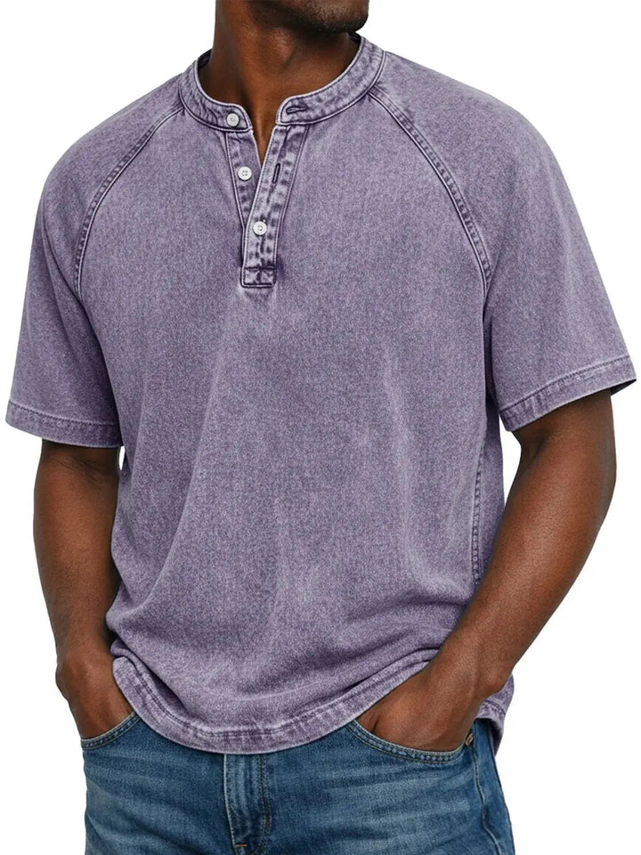 Man draagt het gewassen Henley-shirt met raglanmouwen in paars, met handen in de zakken van een spijkerbroek. 1