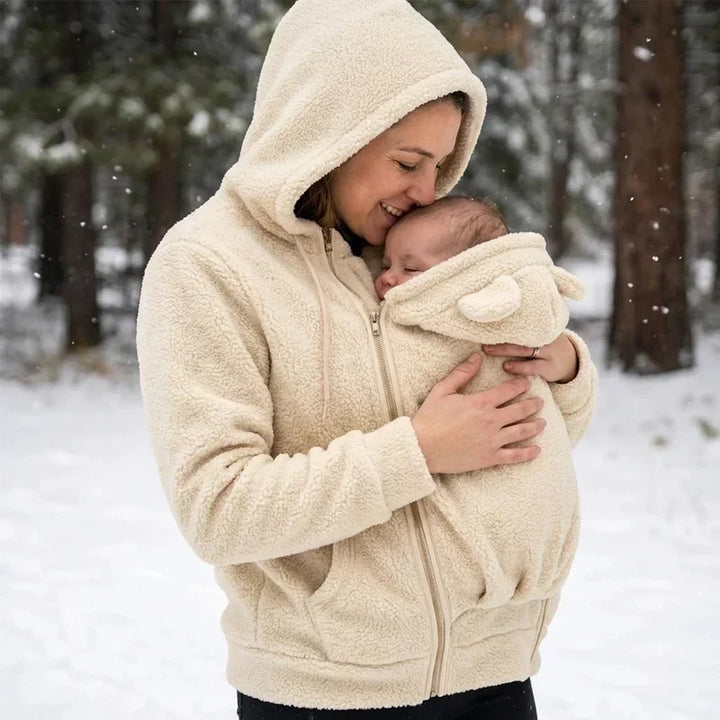 Moeder in beige sherpa hoodie met capuchon op, kust haar baby in het draagpaneel terwijl het sneeuwt in het bos. 8