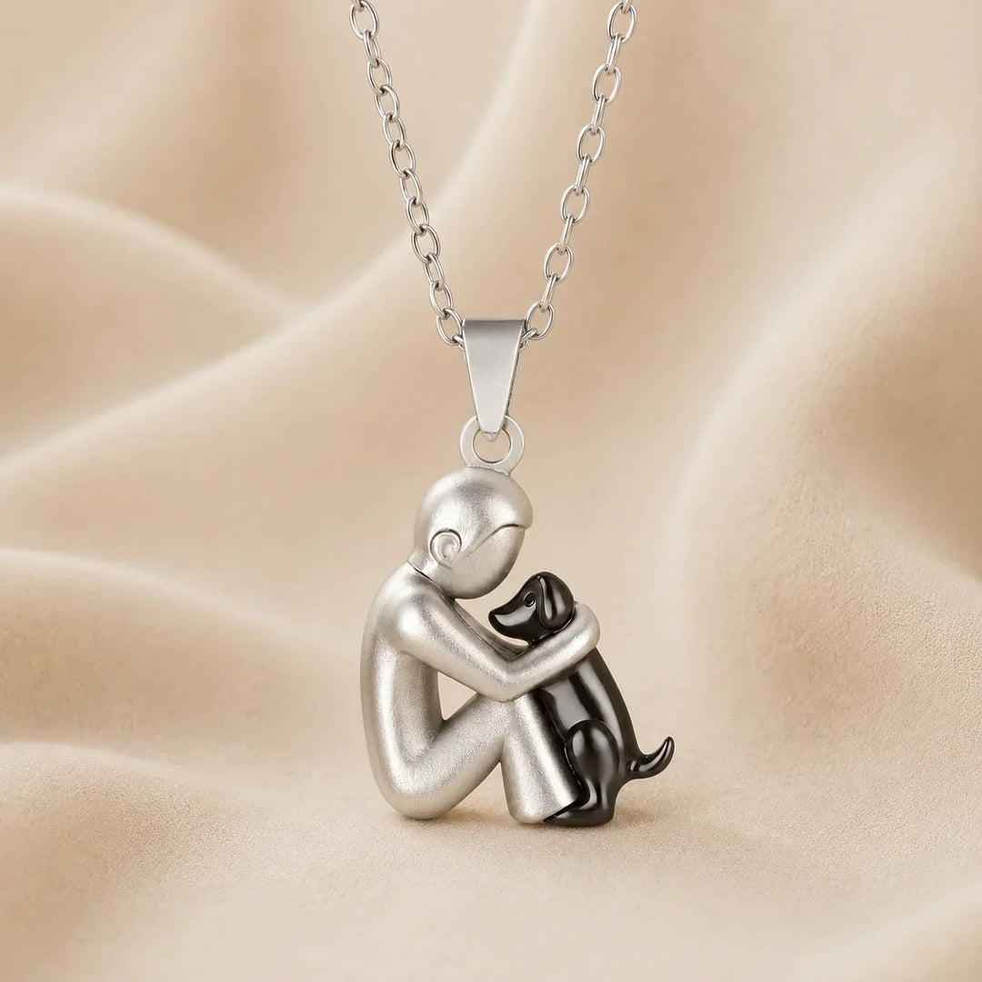 Hanger van een mat zilverkleurige figuur die een kleine zwarte hond vasthoudt, aan een zilverkleurige ketting op beige stof. 7