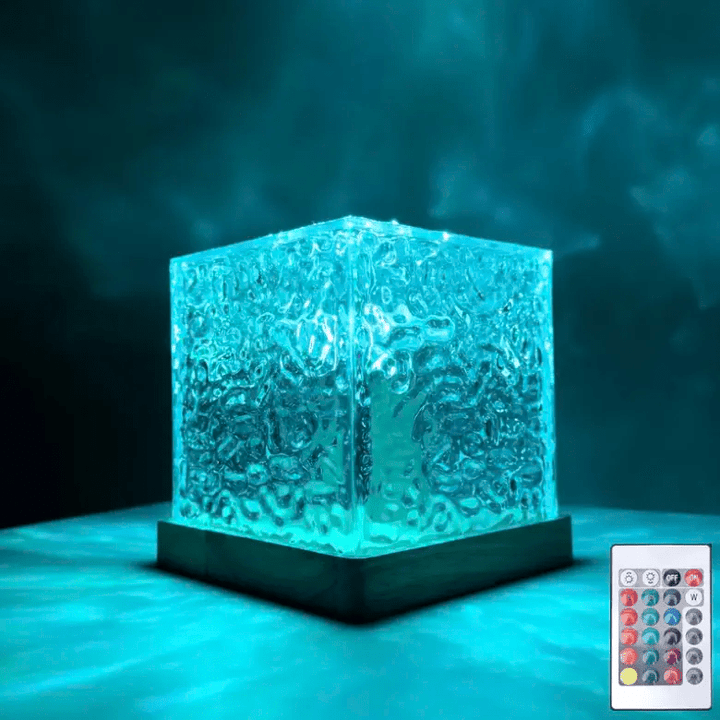 Doorzichtige kubuslamp met golvend oppervlak op houten voet, turquoise verlicht, met een afstandsbediening in beeld in de hoek. 1