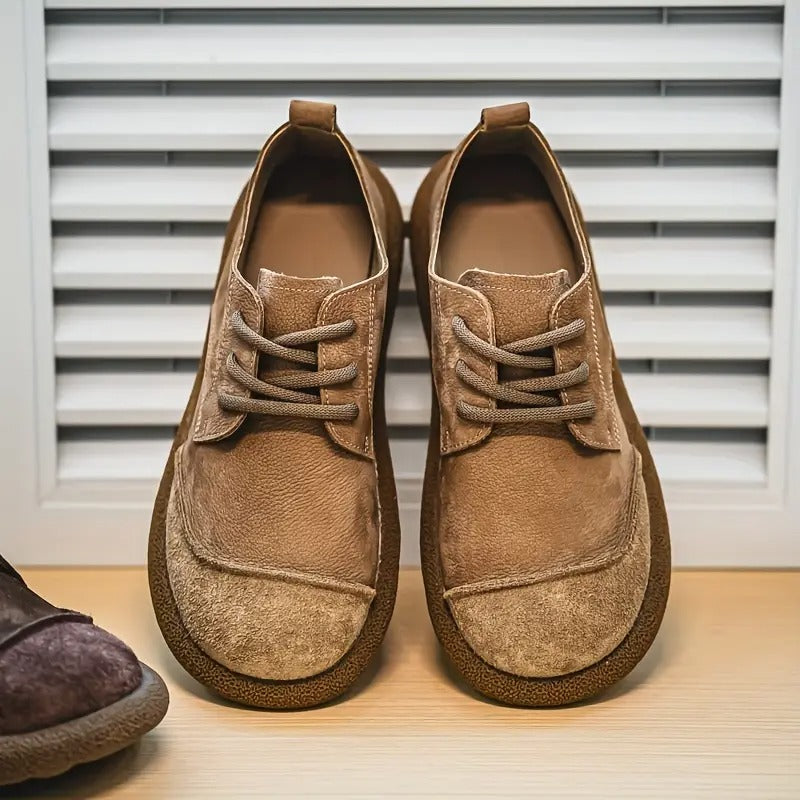Paar bruine suède veterschoenen met brede ronde neus en rubberen zool, van voren gezien op een houten plank voor een lamellenwand. 1