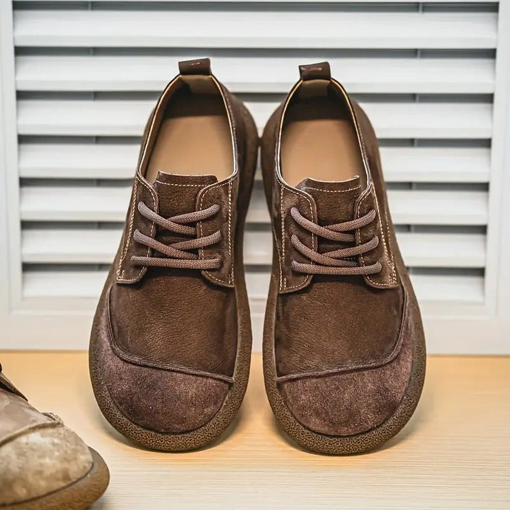 Paar donkerbruine suède veterschoenen met zichtbare stiksels en rubberen zool van voren op een houten plank voor een lamellenwand. 10