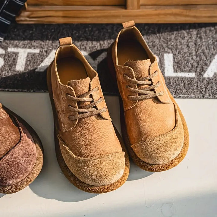 Paar camel suède veterschoenen met brede neus en gekleurde zool, schuin van boven op een lichte ondergrond bij een deurmat. 2