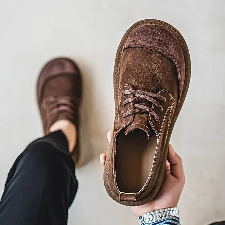 Hand houdt een donkerbruine veterschoen omhoog met zicht op de binnenkant, terwijl de andere schoen aan de voet bij een zwarte broek staat. 7