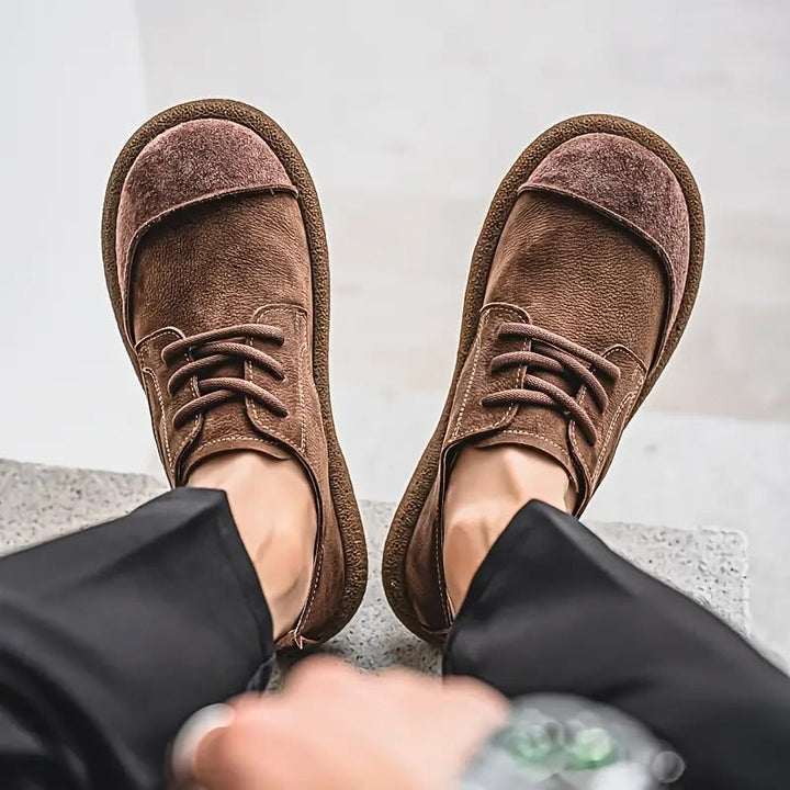 Bovenaanzicht van donkerbruine veterschoenen aan de voeten van een persoon in zwarte broek, met zichtbare contrasterende stiksels en brede neus. 8