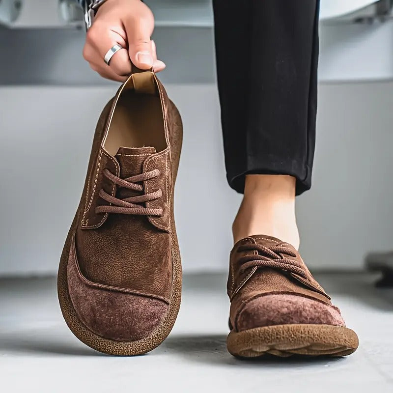 Man houdt één donkerbruine suède veterschoen omhoog aan het lipje, de andere schoen draagt hij aan zijn voet bij een zwarte broek. 6