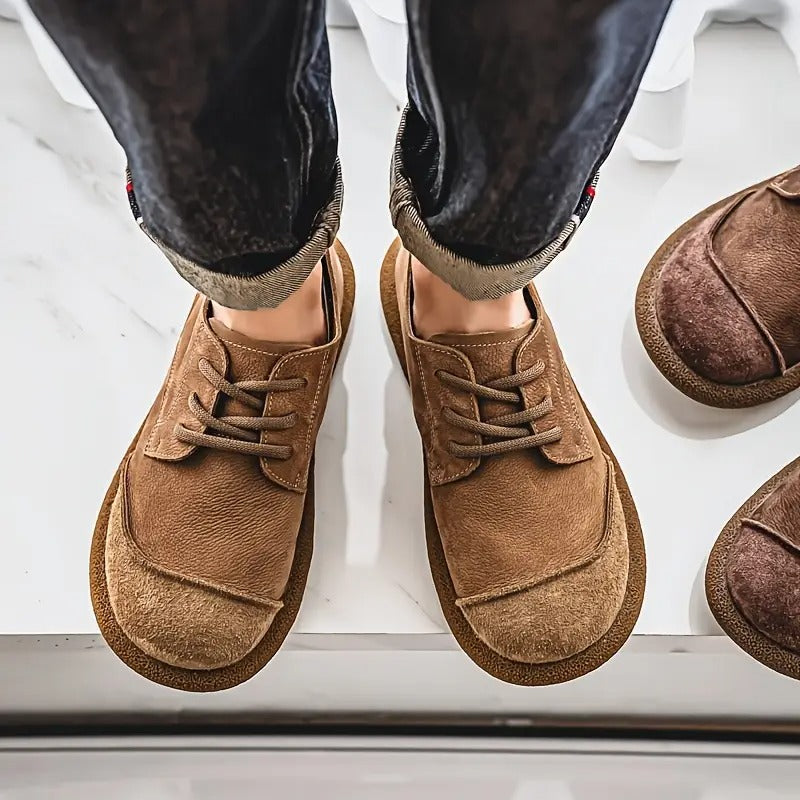 Bovenaanzicht van bruine veterschoenen aan de voeten van een man in opgerolde spijkerbroek, met een donkerder paar ernaast op marmer. 4
