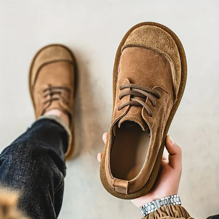 Hand toont een bruine veterschoen van boven met de binnenkant zichtbaar, terwijl de andere schoen aan de voet bij een spijkerbroek zit. 5