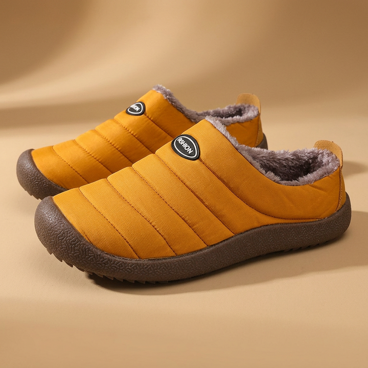 Paar mosterdgele pantoffels met bruine pluche voering en stevige bruine zool op een zandkleurige achtergrond 20