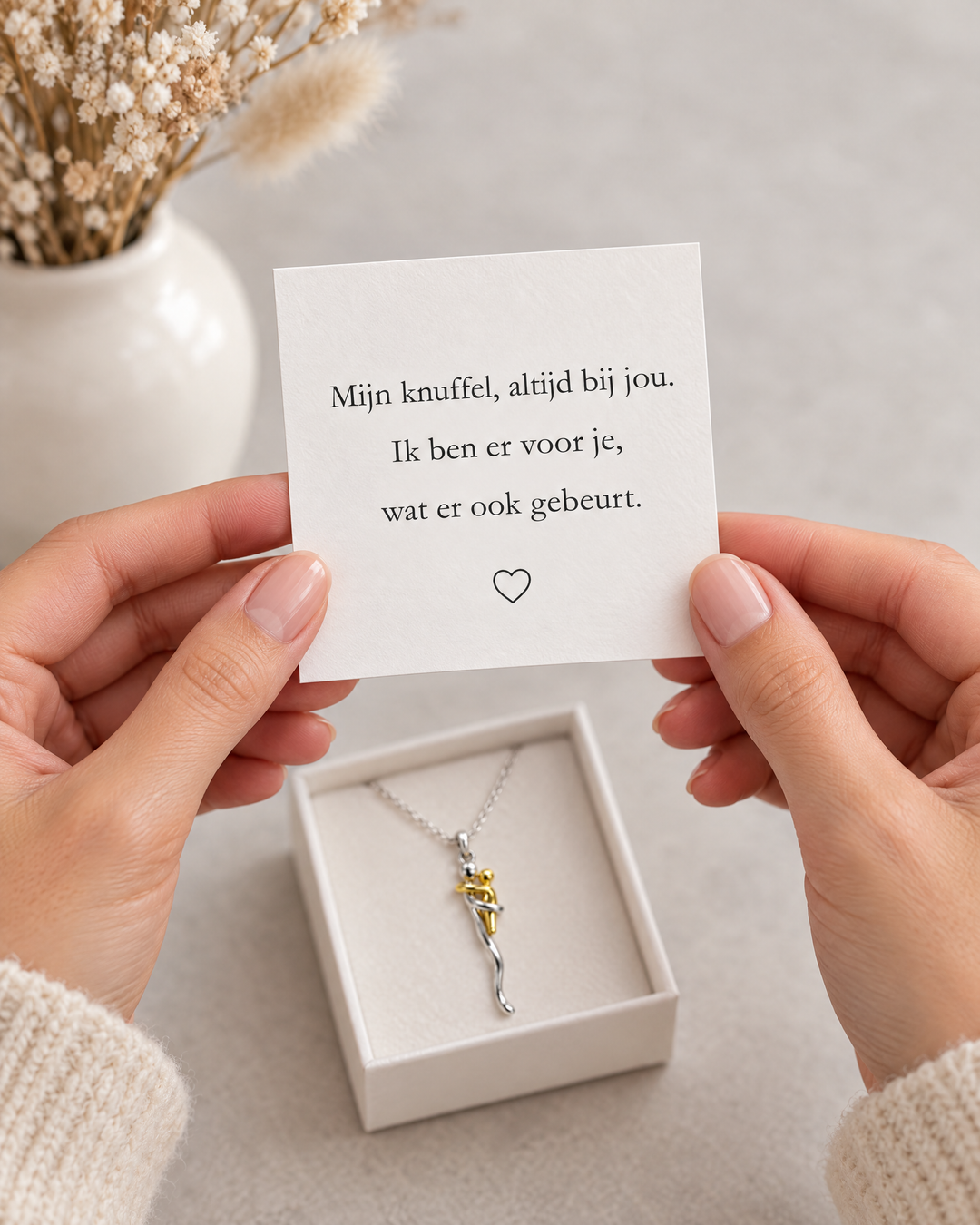 Twee handen houden een wit kaartje met een liefdevolle tekst en hartje vast boven een doosje met de ketting. 6