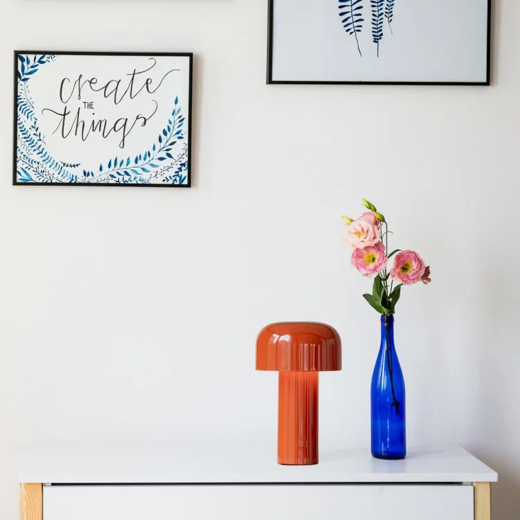 Oranje paddenstoellamp op een witte commode naast een blauwe fles met roze bloemen onder ingelijste prints 19