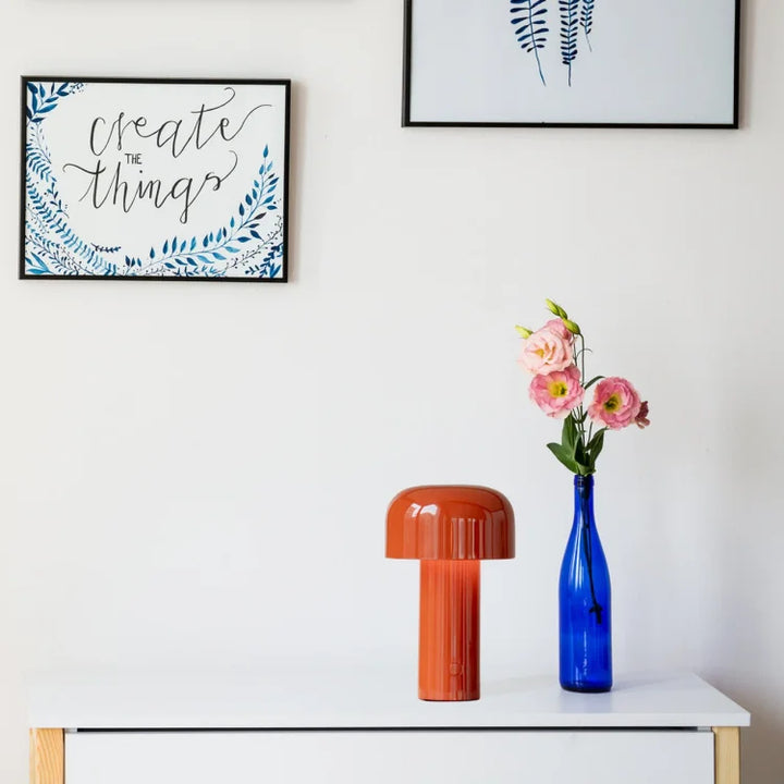 Oranje paddenstoellamp op een witte commode naast een blauwe fles met roze bloemen onder ingelijste prints 19