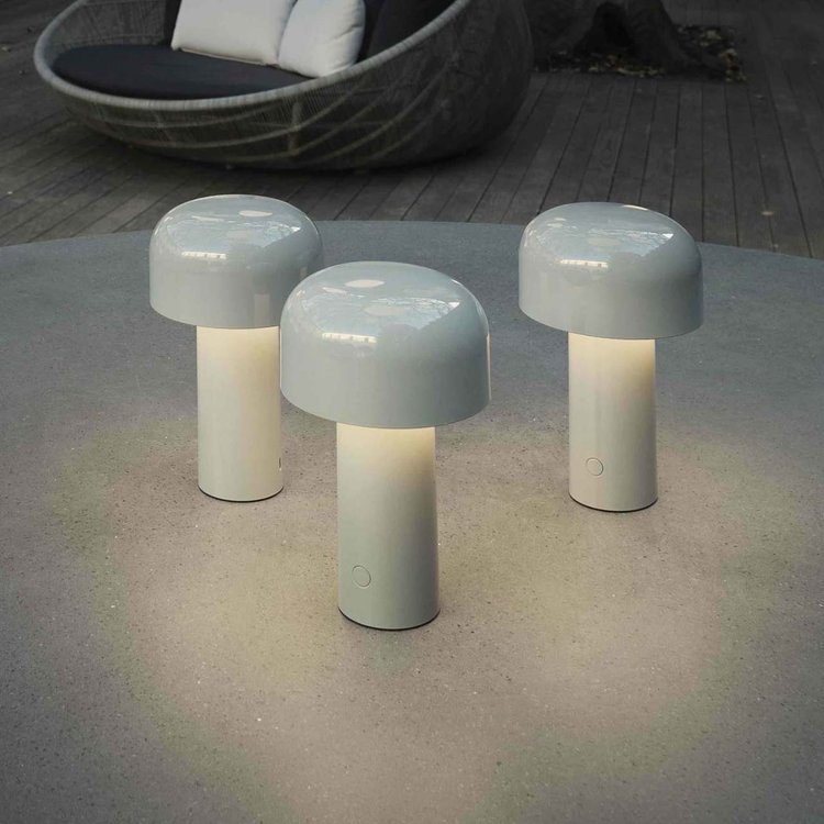 Drie lichte paddenstoelvormige tafellampen branden buiten op een terrastafel, met een rotan zitelement op de achtergrond 6