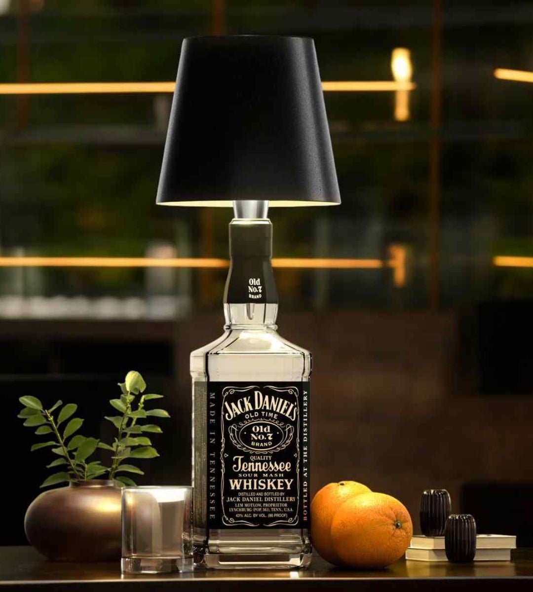 Zwarte lampenkap op een vierkante whiskyfles, naast een glas, een plantje en sinaasappels op een tafel. 2