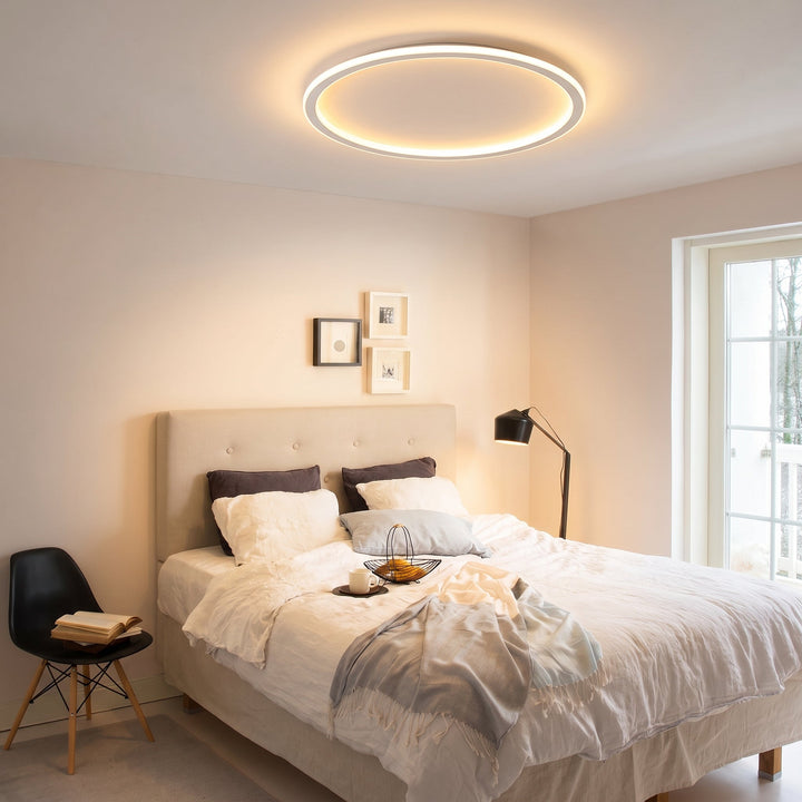 Plafondlamp met lichtgevende ring boven een slaapkamer met opgemaakt tweepersoonsbed, vloerlamp en een zwarte stoel. 9
