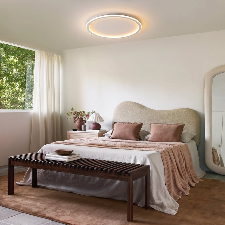 Ringvormige plafondlamp in een lichte slaapkamer met bouclé hoofdbord, roze beddengoed, houten bankje en spiegel. 11