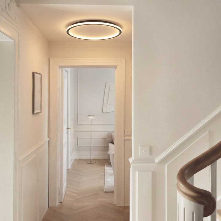 Ronde plafondlamp brandend in een lichte hal met witte lijstwerken, een parketvloer en een trapleuning van hout. 5