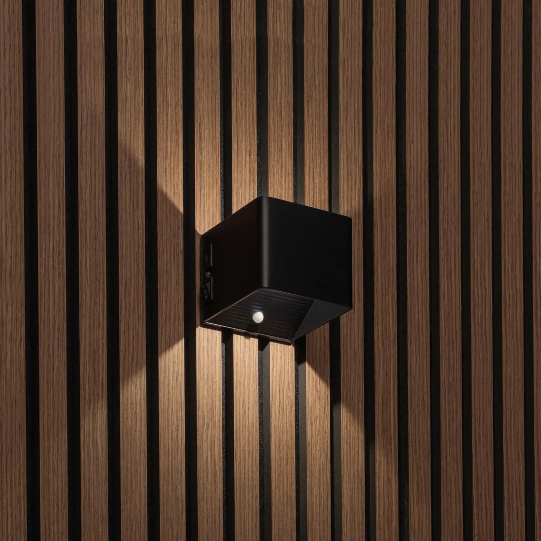 Zwarte kubusvormige wandlamp op houten lamellenwand, schuin gezien met bewegingssensor, schakelaar en lichtstralen omhoog en 1