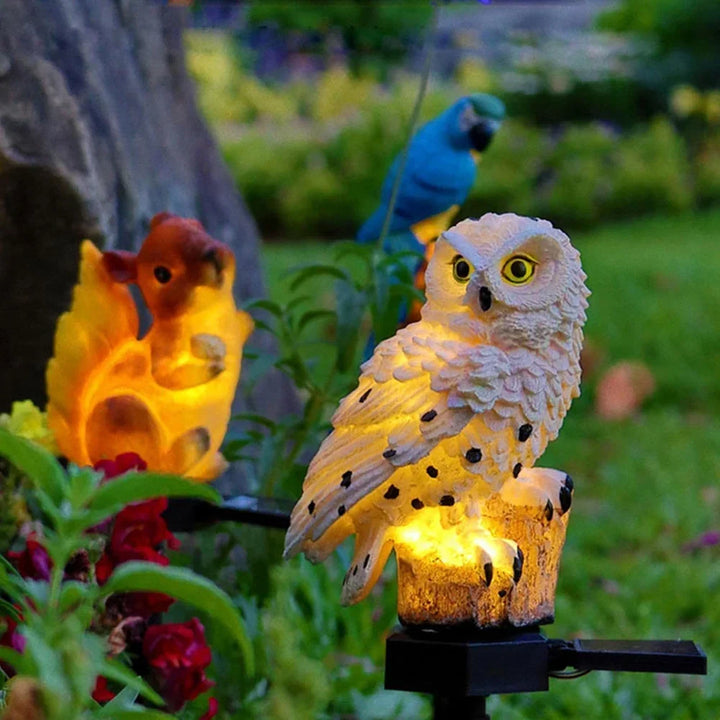Verlichte witte uilenlamp in een tuin naast een oranje eekhoornlamp, met een blauwe papegaai op de achtergrond. 12