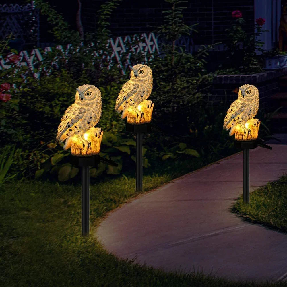 Drie brandende uilenlampen op prikstokken langs een tuinpad in het donker, omgeven door struiken en bloemen. 3