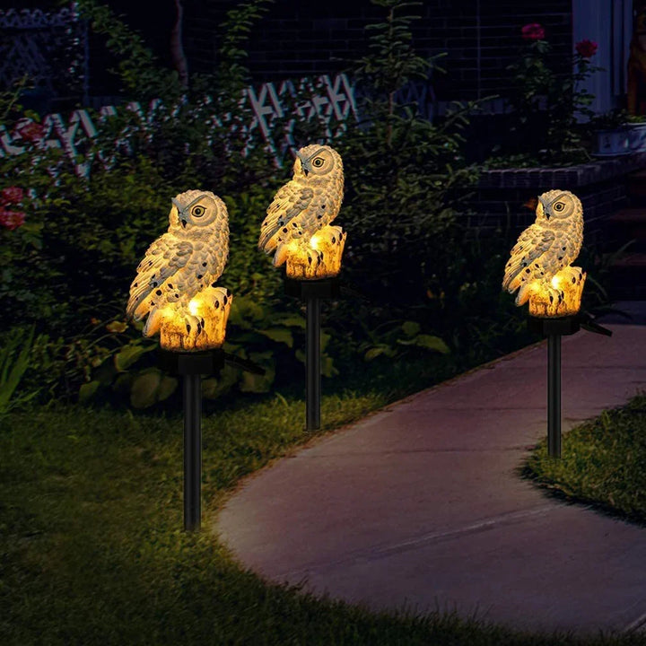 Drie brandende uilenlampen op prikstokken langs een tuinpad in het donker, omgeven door struiken en bloemen. 3