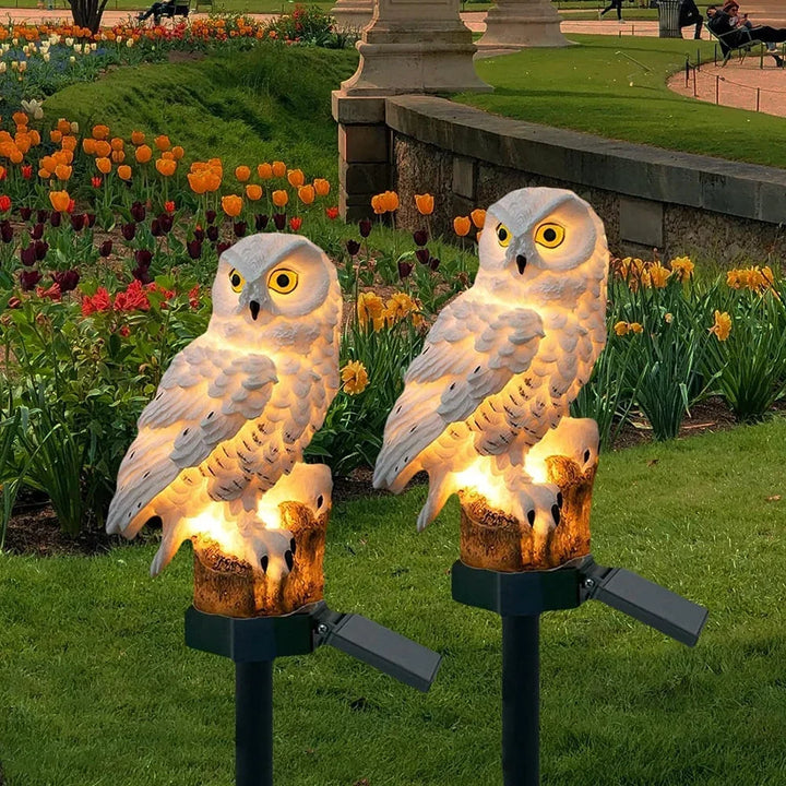 Twee witte uilenlampen op prikstokken met zonnepaneel, brandend in een tuin met oranje tulpen en gele narcissen. 1