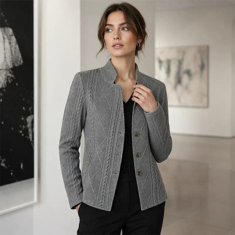 Vrouw in een galerie in een grijze blazer met kabelstructuur, zwarte top en zwarte broek, hand bij de kraag. 11