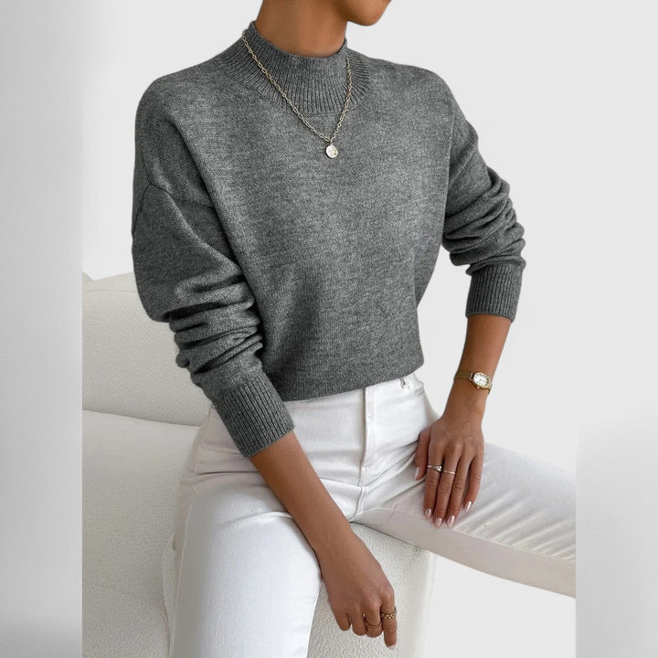 Model draagt de trui in gemêleerd grijs met ribkraag en lange mouwen, met witte broek en een dunne ketting. 12