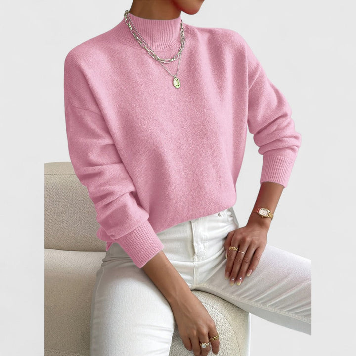 Model draagt de trui in zacht roze met ribboord aan kraag en mouwen, met witte broek en zilveren kettingen. 5
