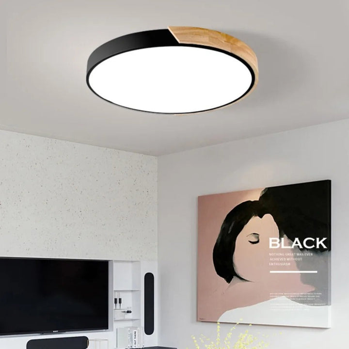 Zwarte ronde plafondlamp met houten accent aan de rand, brandend boven een woonkamer met tv en kunstwerk aan de muur. 1