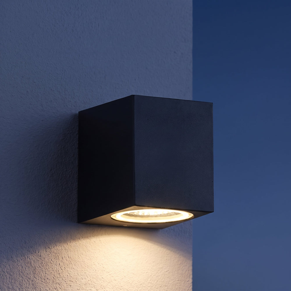 Zwarte kubuslamp aan een gestuukte muur in de schemering, met warm licht naar beneden 2