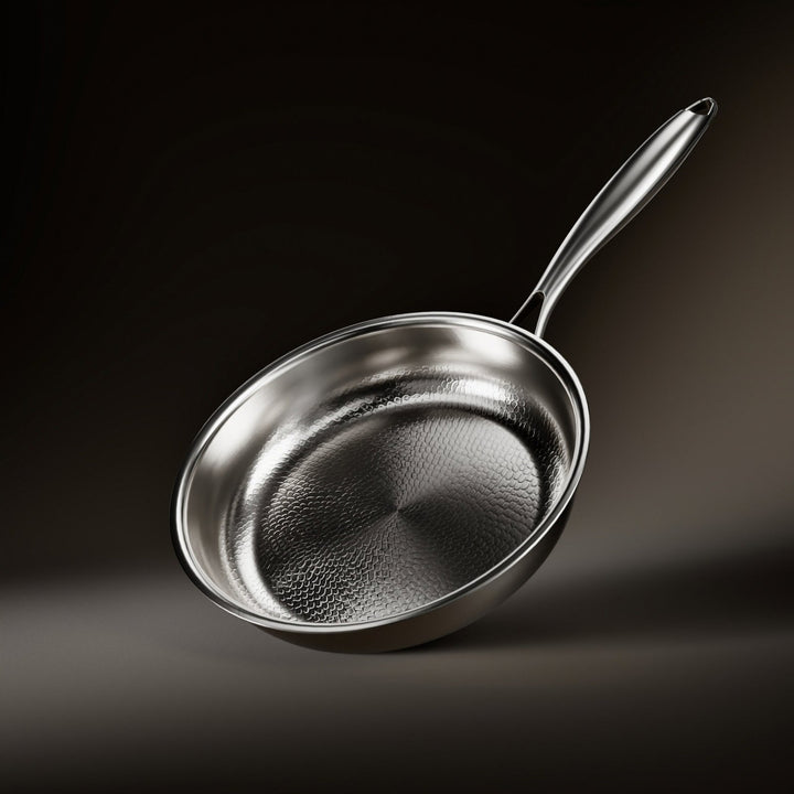 Schuin bovenaanzicht van de koekenpan met gehamerd patroon in het zilverkleurige binnenoppervlak en een lange metalen steel, tegen een donkere achtergrond. 1