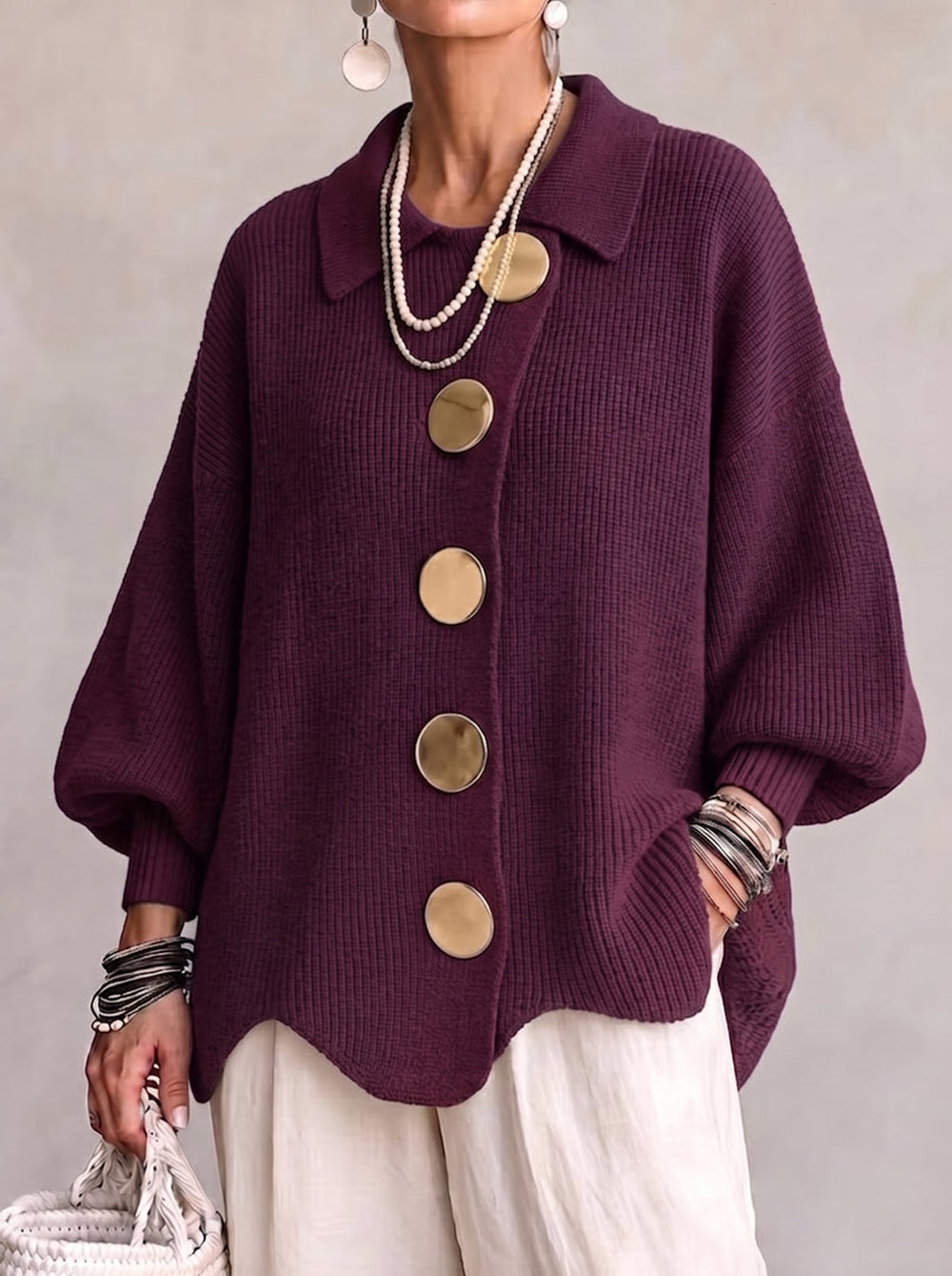 Het gebreide vest in donker aubergine met grote goudkleurige knopen, gedragen met parelkettingen, wijde witte broek en rieten tas. 2